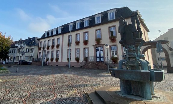 Seitlicher Blick auf das Saarwellinger Rathaus. Im vorderen, rechten Bildbereich steht ein Brunnen.