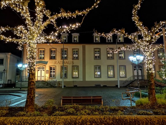 Blick auf das Saarwellinger Rathaus bei Nacht. In den Fenstern hängt Weihnachtsbeleuchtung. Im Vordergrund mit Lichterketten geschmückte Bäume.