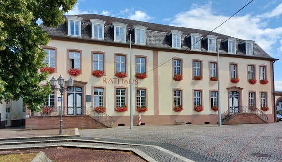 Außenansicht Front des Rathauses Saarwellingen. Blauer Himmel mit wenigen Schleierwolken, rot blühende Geranienblumen auf allen Fensterbänken des Rathauses. Im linken Bildrand sieht man die grünen Blätter eines Baumes.